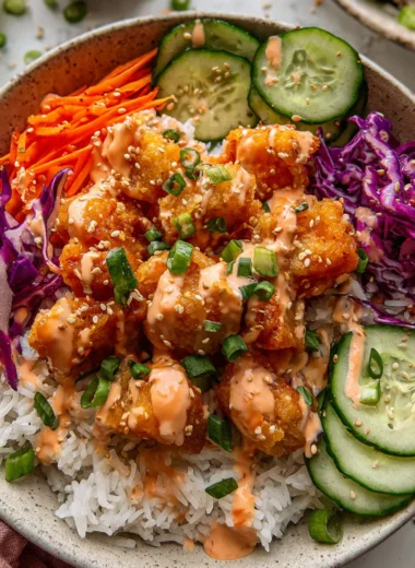 Bang bang chicken bowl with crispy sauced chicken, fluffy rice, cucumber, carrots, purple cabbage, and sesame seeds in a wide ceramic bowl