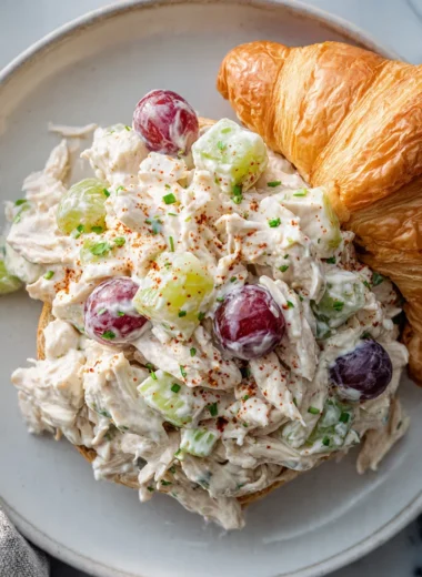 Close-up of classic protein chicken salad with shredded chicken, grapes, and celery served on a white plate next to a croissant.
