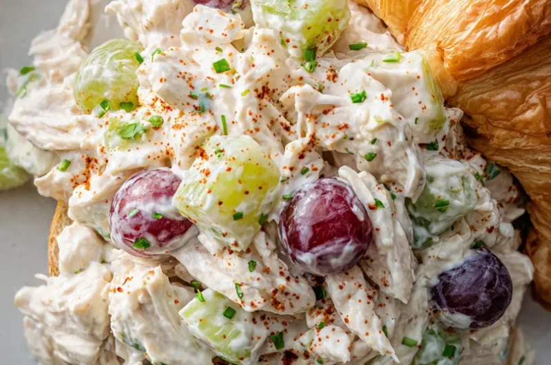 Close-up of classic protein chicken salad with shredded chicken, grapes, and celery served on a white plate next to a croissant.