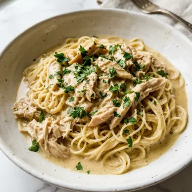 Crock Pot Angel Chicken served over angel hair pasta in a white ceramic bowl with creamy golden sauce and fresh parsley