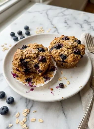 Close-up of high-protein oatmeal blueberry muffins with golden tops and jammy blueberry pockets on a white ceramic plate