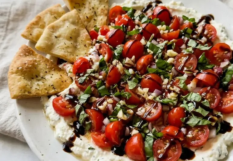 Bruschetta Dip on a white plate with tomatoes, basil, balsamic glaze, and pita chips ready for dipping