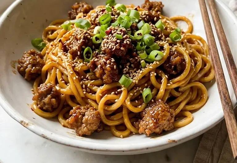 Close-up of sticky beef noodles in a white bowl topped with spring onions and sesame seeds