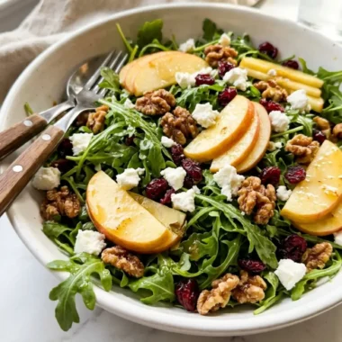 apple arugula salad in a white ceramic bowl with walnuts, goat cheese, and cranberries