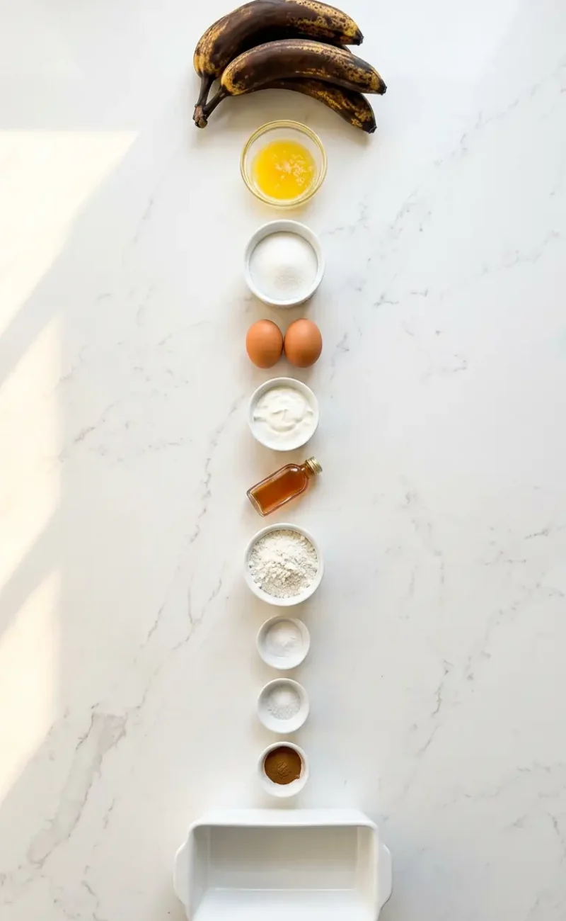 Exploded view of all 11 ingredients for banana bread recipe arranged vertically on white marble