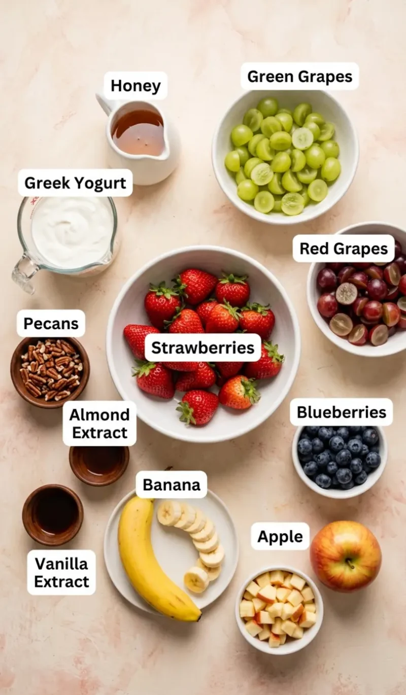 Labelled overhead flatlay of all 11 ingredients for breakfast fruit salad on a cream and blush plaster surface, each ingredient named
