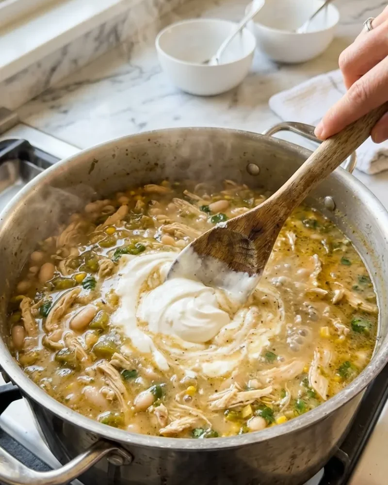 Sour cream being stirred into a pot of creamy white chicken chili with steam rising and visible beans and chicken