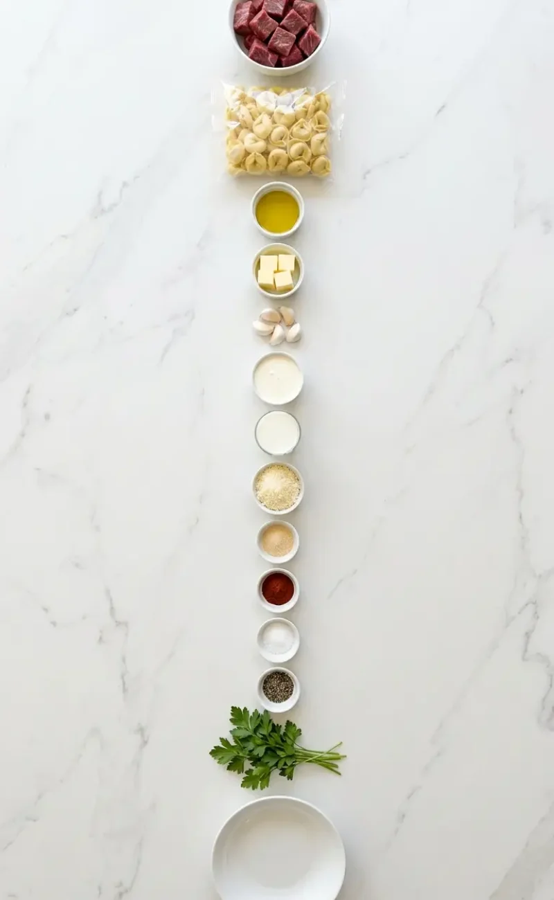 Exploded view of all 13 ingredients for garlic steak tortellini arranged vertically on white marble