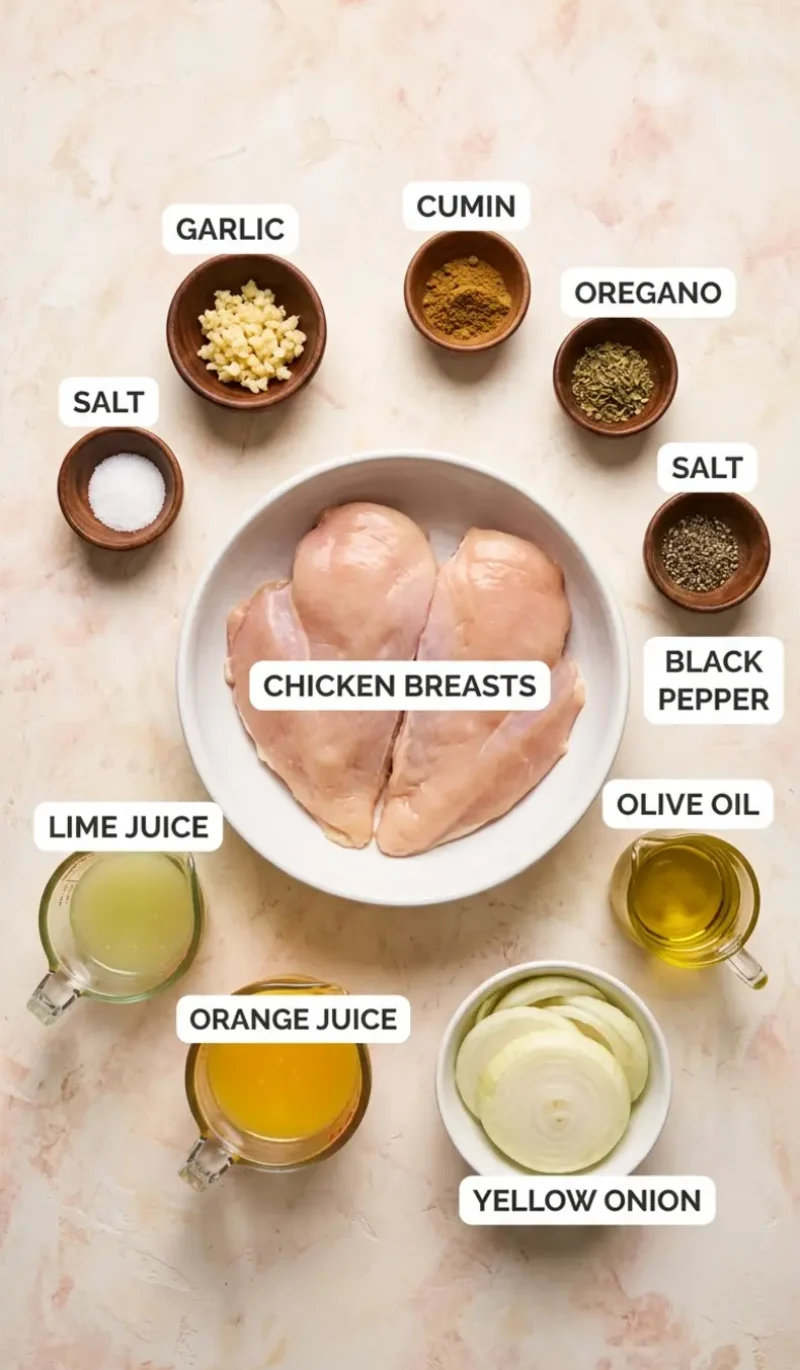 Labelled overhead flatlay of all 10 ingredients for Pollo a la Plancha (Cuban Chicken) on a cream and blush plaster surface, each ingredient named