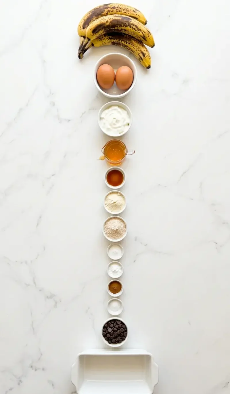 Exploded view of all 12 ingredients for protein-packed banana bread arranged vertically on white marble
